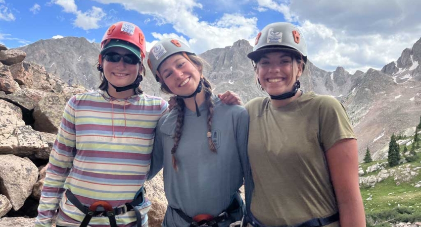 three students wearing helmets smile for a photo in front of a cast mountainous landscape